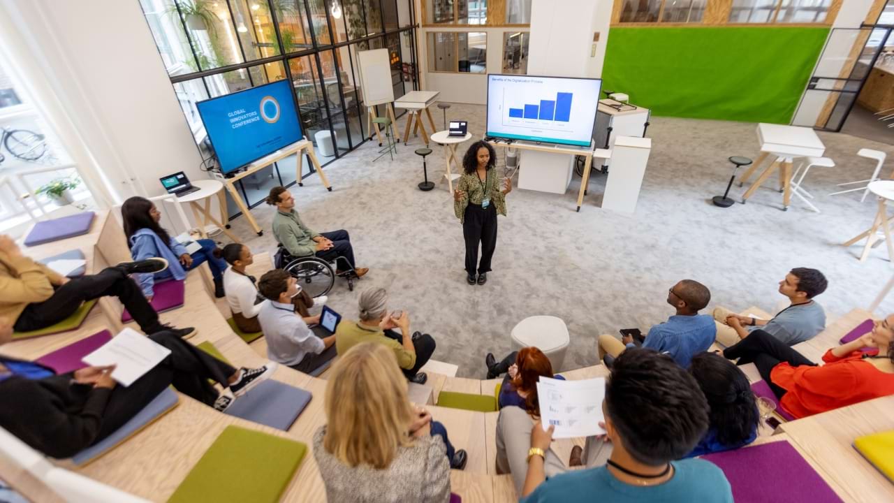 High-angle view of a diverse group of stakeholders in a project kickoff meeting, seated in a modern amphitheater–style workspace with plans and laptops spread before them